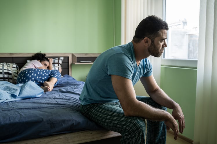 An unhappy man sitting on the edge of his bed at home, while his wife lies unhappily in the bed.