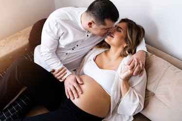 A man kissing a pregnant woman on a couch, with a hand on her pregnant belly.