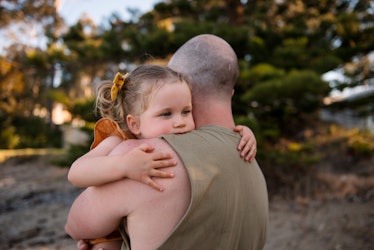 A grumpy young girl hugs her father while they stand outside on a beach.