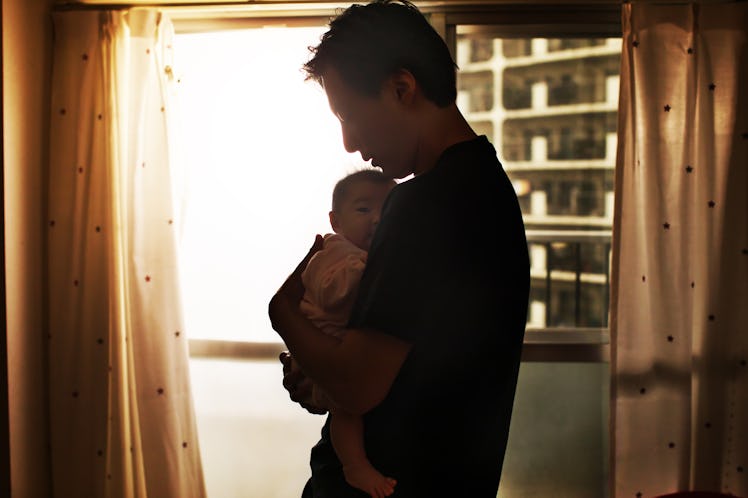 A new dad with postpartum depression holding his baby in a dimly lit room, by a window.