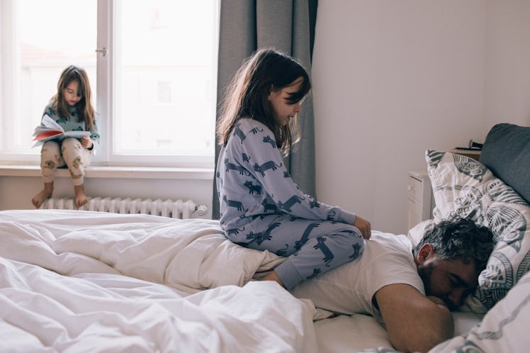 A girl sitting on her dad's back as he sleeps in bed, as another girl sits to the side, reading a bo...