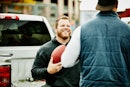 一个面带微笑的男人拿着足球，在一辆小货车后面和朋友聊天