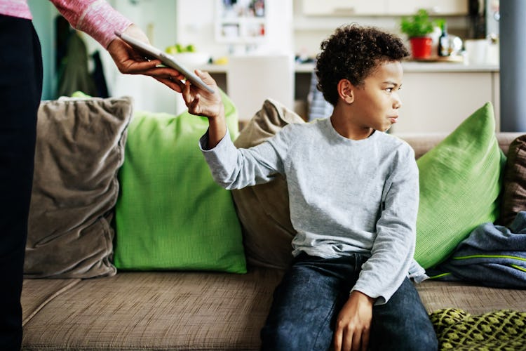 A parent taking away a child's tablet as punishment as the kid sits on a couch and frowns.