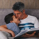 A dad reading a book to his son on a couch takes a break to kiss his son on the nose.