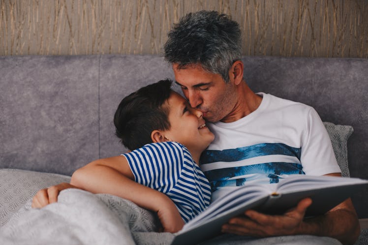 A dad reading a book to his son on a couch takes a break to kiss his son on the nose.