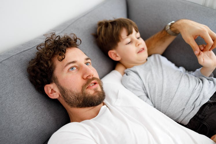 A dad with his arm around his son's shoulders, leaning back on a couch.