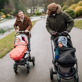 Two dads pushing their babies in strollers down a path in a park.