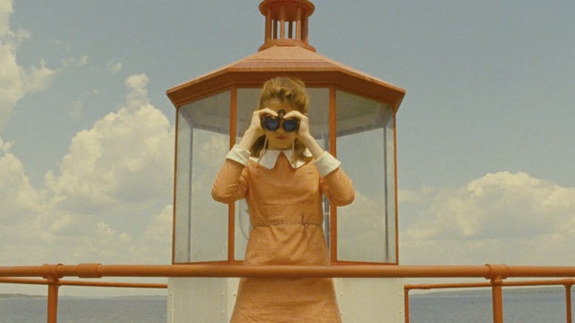 A woman wears a yellow dress with white collars and looks into binoculars.