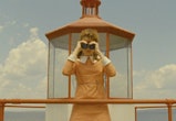 A woman wears a yellow dress with white collars and looks into binoculars.