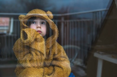 A child in a bear onesie staring out a window, depressed.