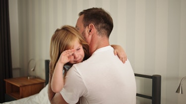 A dad comforts his child, who is crying into his shoulder as they sit on a bed.