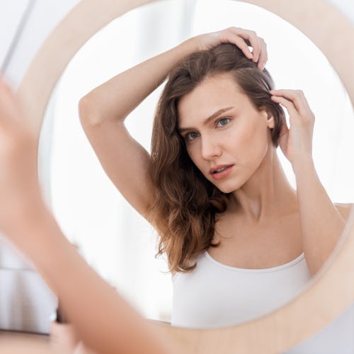 young woman touching her scalp while looking in the mirror