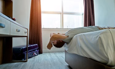 A man lying with his head off the bed, while his unpacked suitcase sits in a corner of the bedroom.