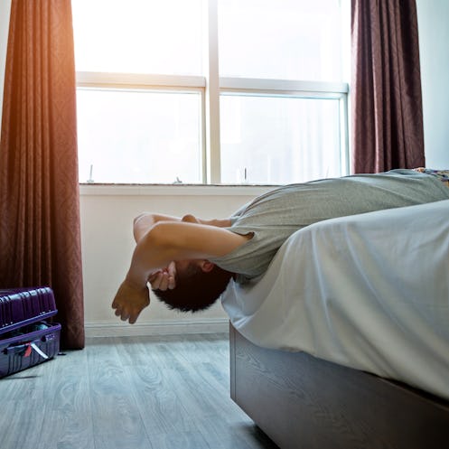 A man lying with his head off the bed, while his unpacked suitcase sits in a corner of the bedroom.