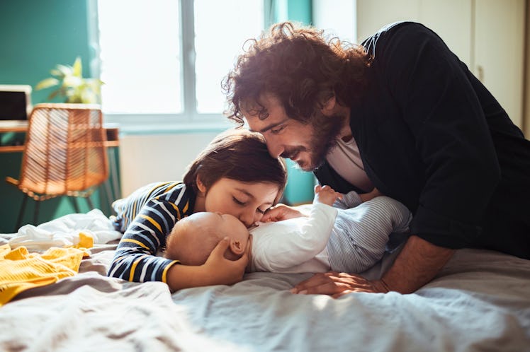 A dad talks to his baby, who lies on a bed, while his older daughter kisses the baby.