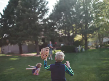 The back of a boy with a baseball glove playing baseball with his family in a backyard