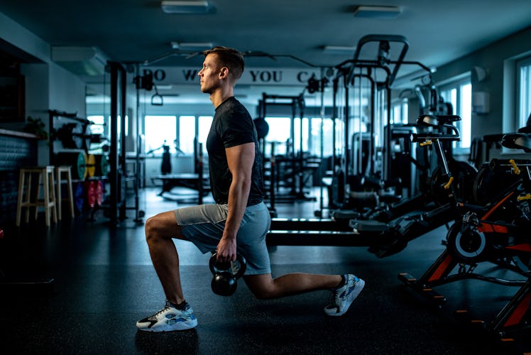 A man doing walking lunges while holding kettlebells in a gym.