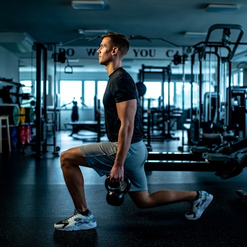 A man doing walking lunges while holding kettlebells in a gym.