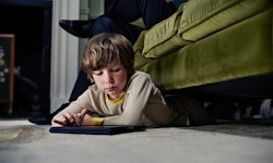 A boy on a tablet while half-lying under a couch that a parent is sitting on.