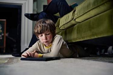 A boy on a tablet while half-lying under a couch that a parent is sitting on.