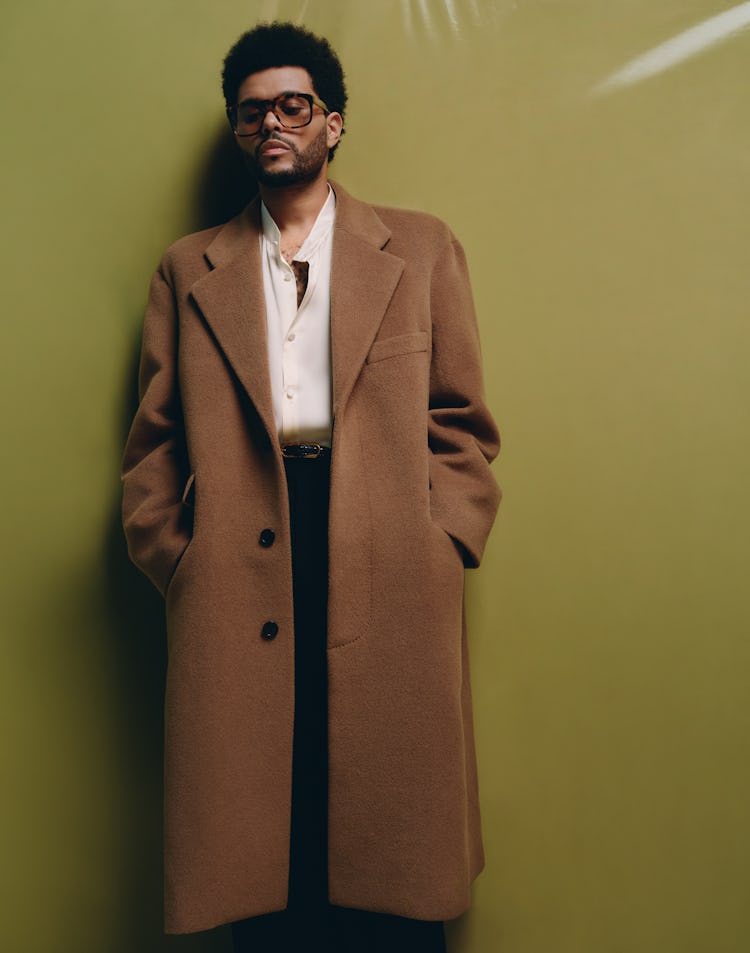 The Weeknd wears a brown coat, sunglasses, black pants, black leather belt and button-down shirt.