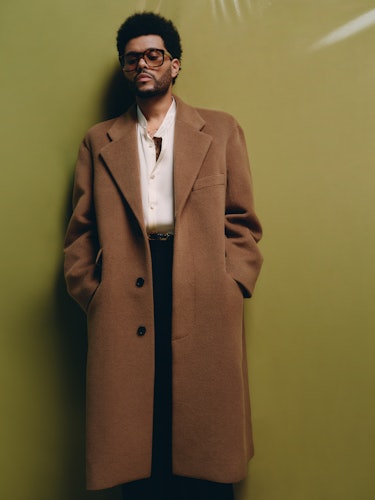 The Weeknd wears a brown coat, sunglasses, black pants, black leather belt and button-down shirt.