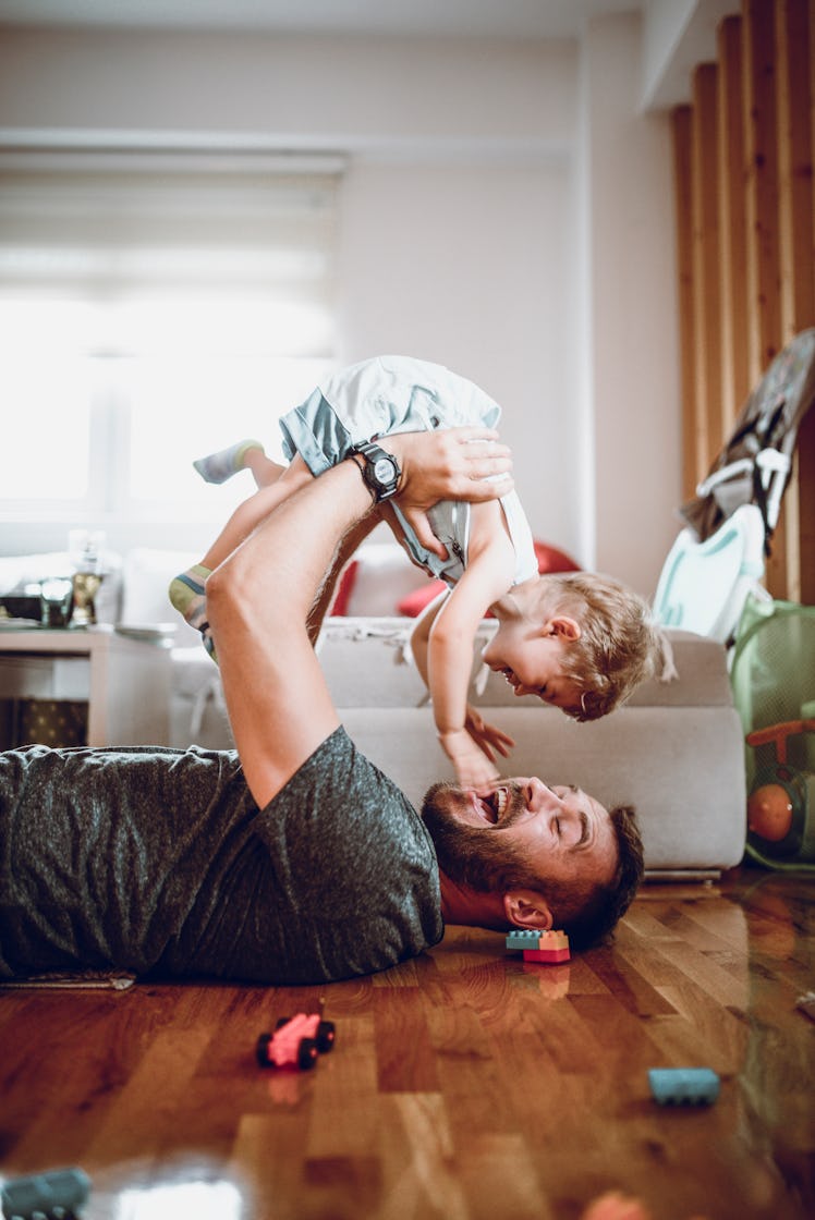 A dad lying on the ground, holding his baby in the air and smiling, with toys surrounding them.