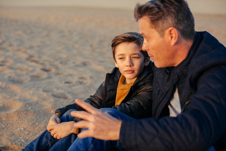 A father and son sitting on a beach, talking.
