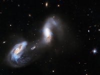 color image of two galaxies, mostly in shades of blue and white, on a black background spangled with...