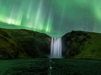 A waterfall flows water over a ridge, and it's illuminated by the auroras in the sky. The auroras lo...