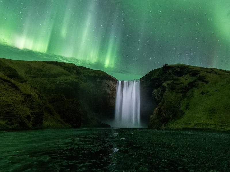 A waterfall flows water over a ridge, and it's illuminated by the auroras in the sky. The auroras lo...