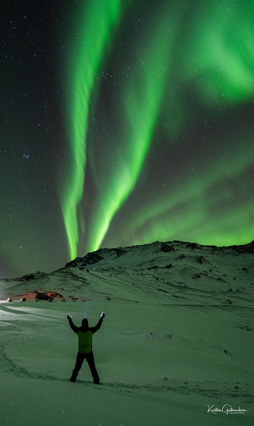 Green bands of aurora spread across the evening sky from behind a snowy ridge. A person stands in a ...