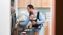 一名男子胸前抱着一个婴儿，用婴儿背带从洗衣机里倒出衣服。