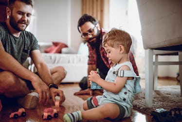 Two dads playing with toys and talking to their toddler on the floor.