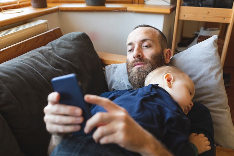 A dad lying on a couch using his phone, while his infant sleeps on his chest.