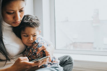 A mom holds her son while sitting by a window, using her cell phone.