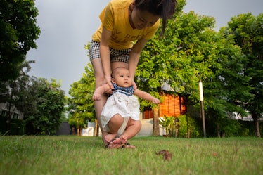 A mom holding a baby above grass as the baby avoids touching the grass by lifting up their legs.