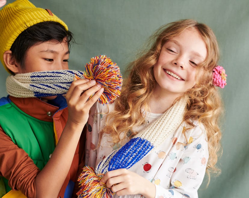 Maisonette Outlet catalog photo of two children in winter wear, playing with pom poms on their scarv...