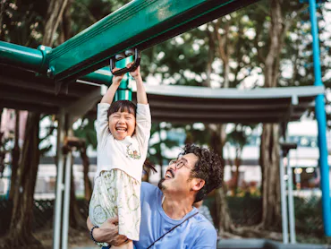 A dad helps his young daughter discover risky play on playground equipment by holding her legs while...