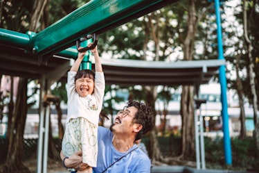 A dad helps his young daughter discover risky play on playground equipment by holding her legs while...