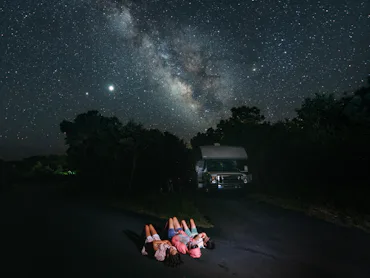 A family on an RV trip stargazes at night, lying on their backs and staring up at the night sky.