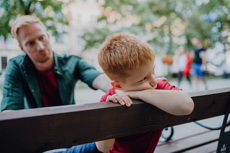 A father consoling his upset son as they sit on a bench in a park.