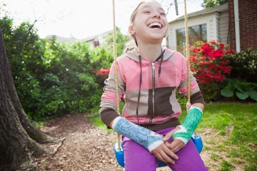 A girl with two broken arms in casts smiles and laughs while sitting on a swing outdoors.