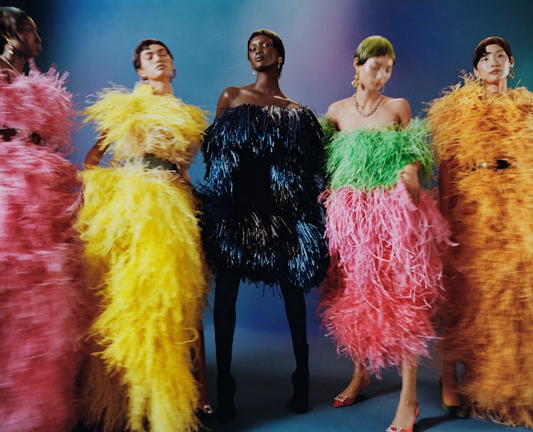 Five models wearing colorful feathered gowns pose against a blue and purple backdrop