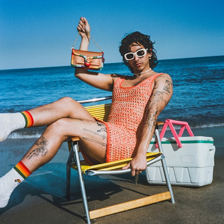 a model holding a coach bag while perched on a beach chair by the ocean