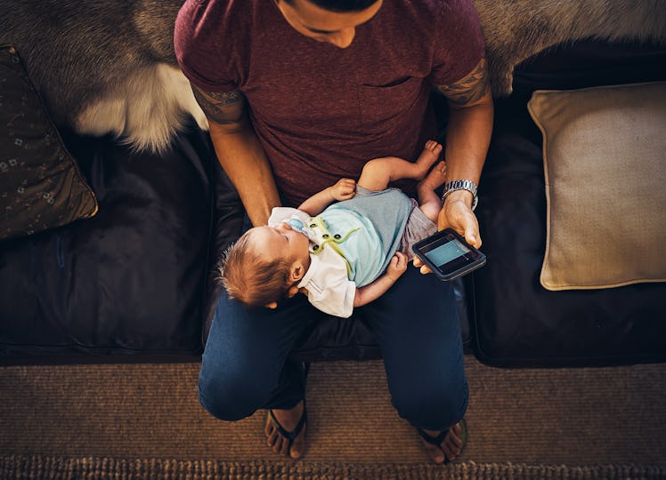A man holding a baby while sitting on a couch and looking at his phone.