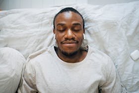A man lying in bed, relaxing with his eyes closed, as he tries autogenic training.