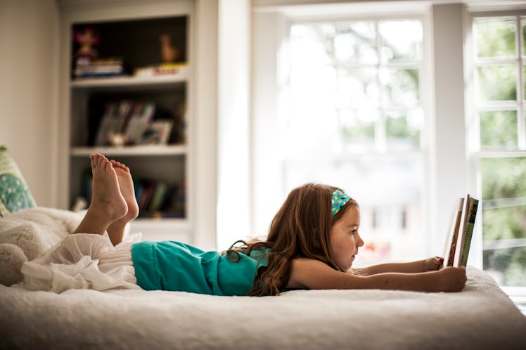 A girl lying on her bed on her stomach, reading a book while home alone.