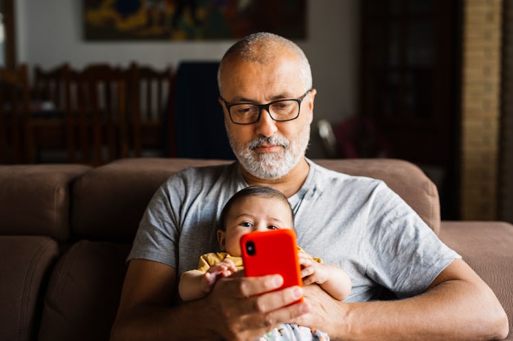 A man holding a baby in his lap while sitting on a couch, staring at his phone with a flat affect.