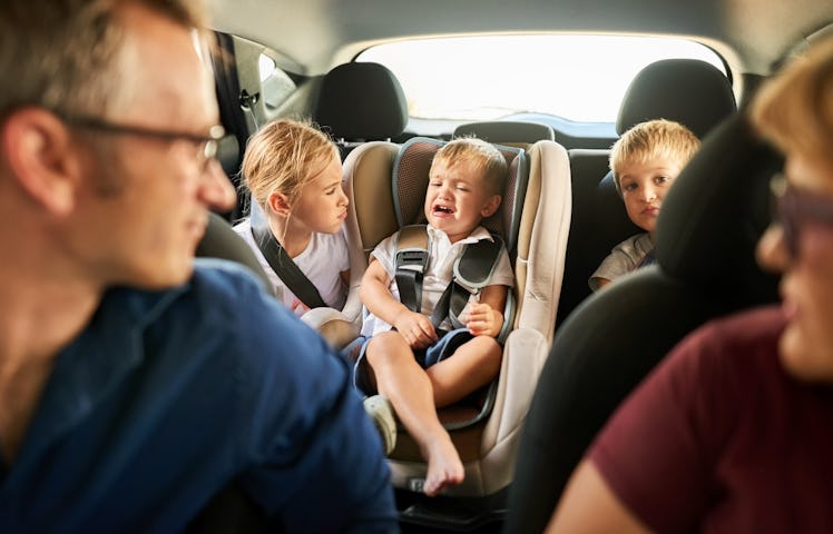 Two parents look at their toddler crying in a car seat, next to his two siblings.